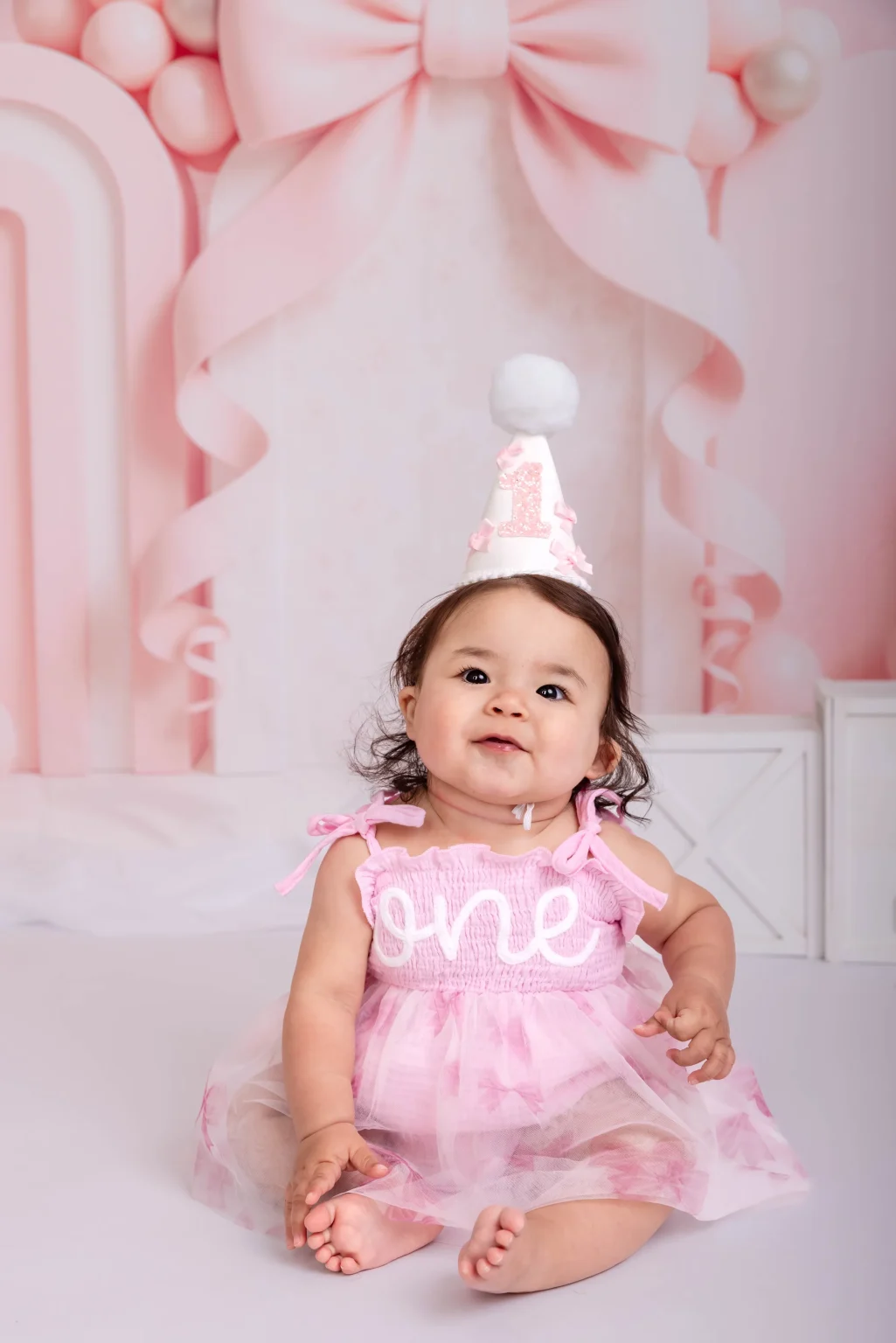 A baby in a pink dress with "one" on it sits smiling, wearing a party hat with the number 1. Pink decorations are in the background.