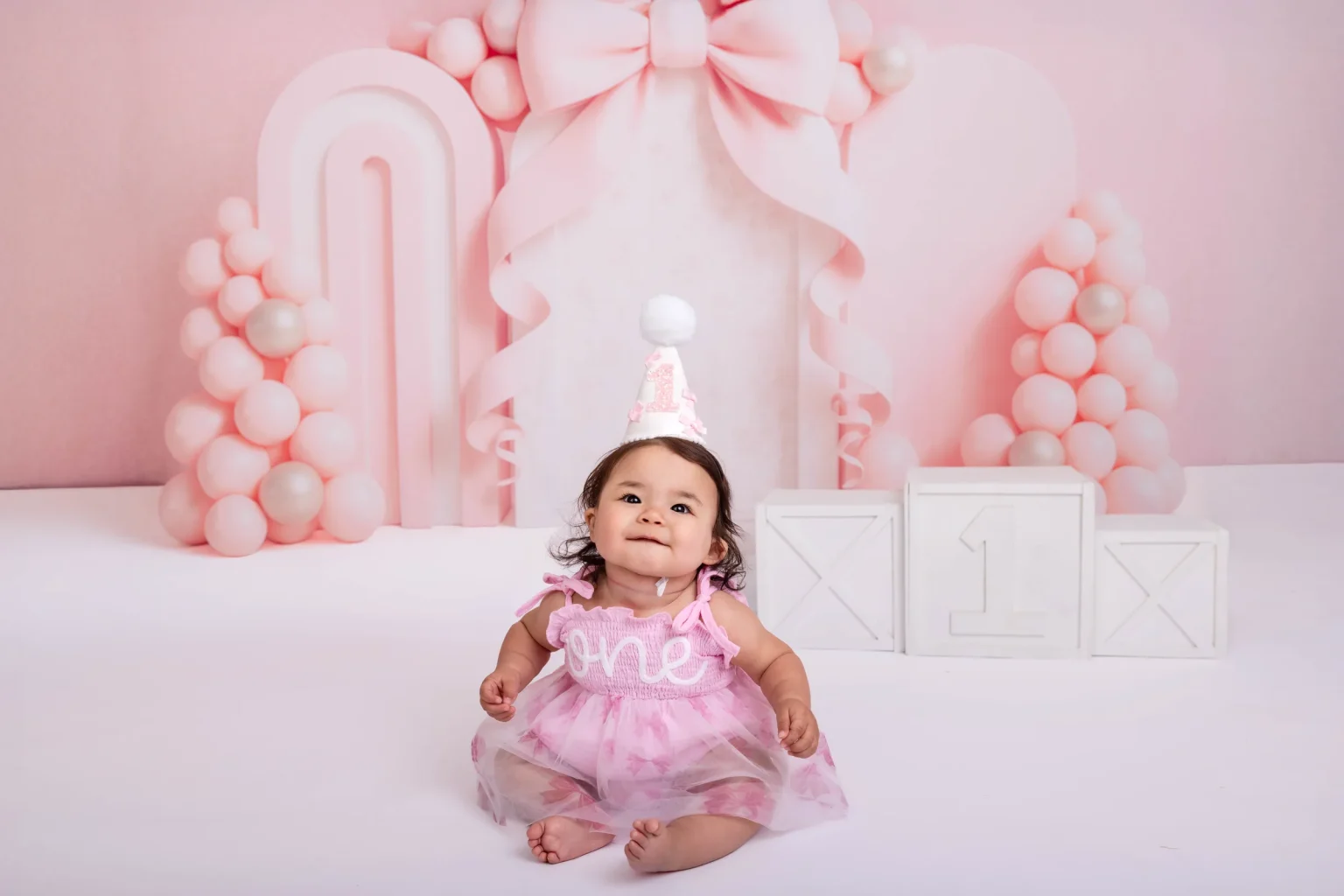 A baby in a pink dress and birthday hat sits in front of pastel pink decorations and blocks marking her first birthday.
