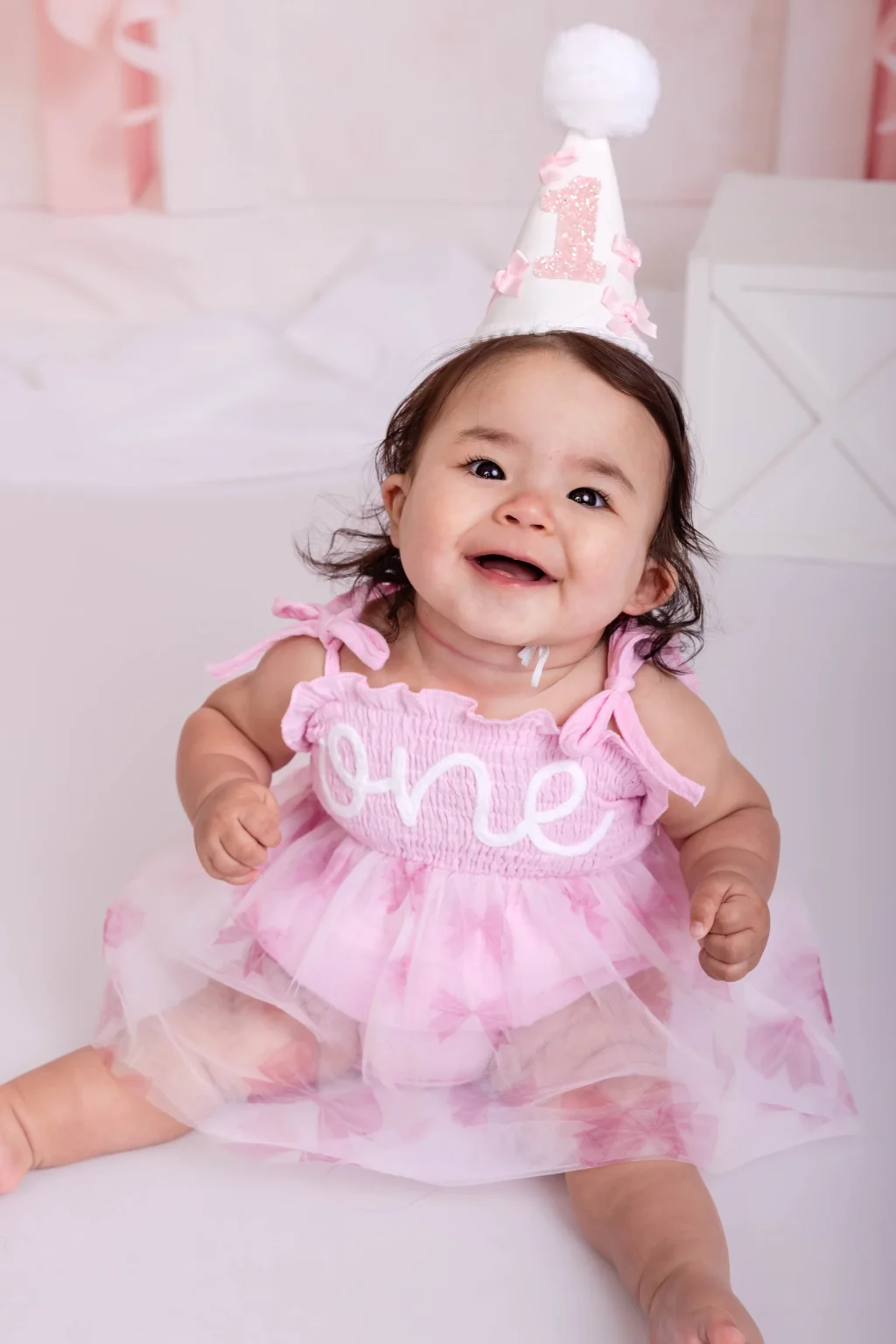 Smiling baby wearing a pink dress with "one" written on it and a birthday hat, sitting indoors, celebrating her first birthday.