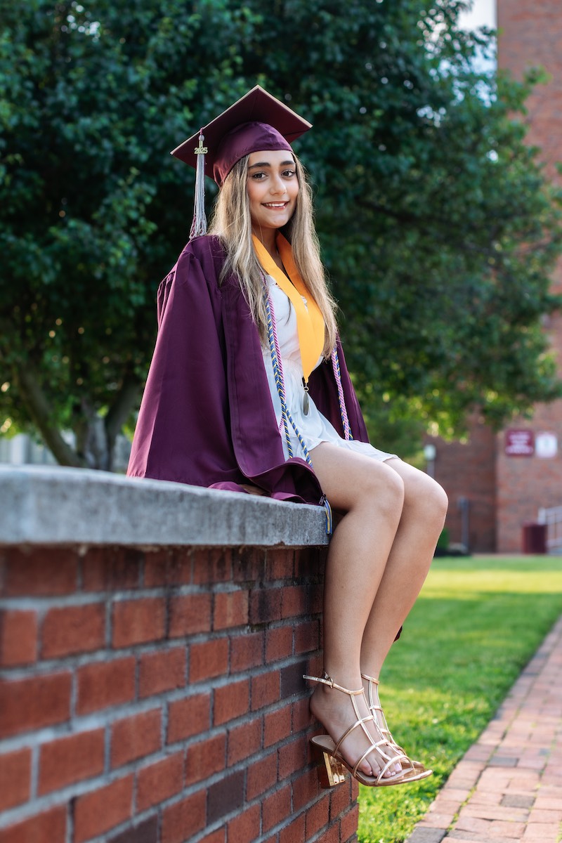 A young woman in a maroon graduation gown and cap sits on a brick wall outdoors, smiling at the camera.