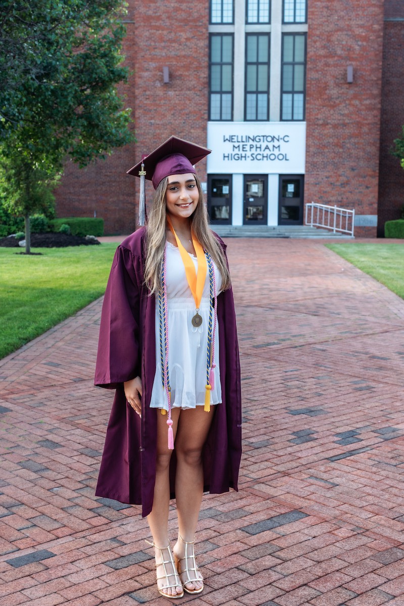 A young woman in a maroon graduation gown and cap stands outside Wellington C. Mepham High School, wearing a white dress, honor cords, and medals.