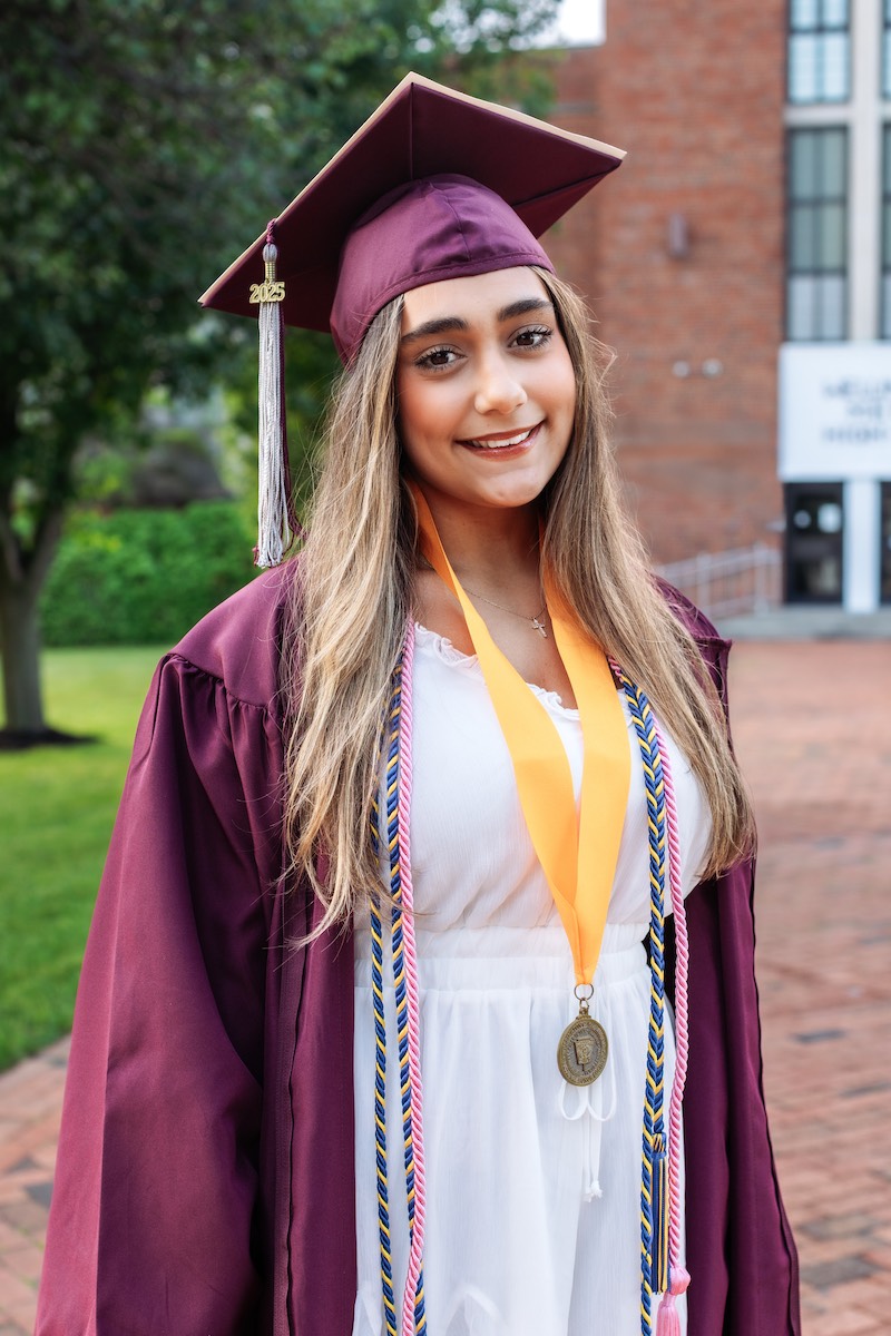 A young woman wearing a maroon graduation cap and gown, honor cords, and a medal, stands outdoors on a brick pathway near a building.