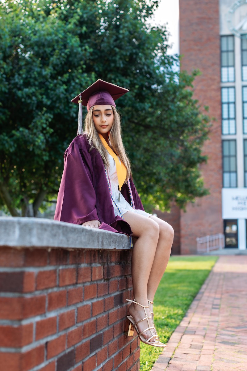 A young woman in a maroon graduation cap and gown sits on a brick wall outdoors, looking down, with a building and trees in the background.