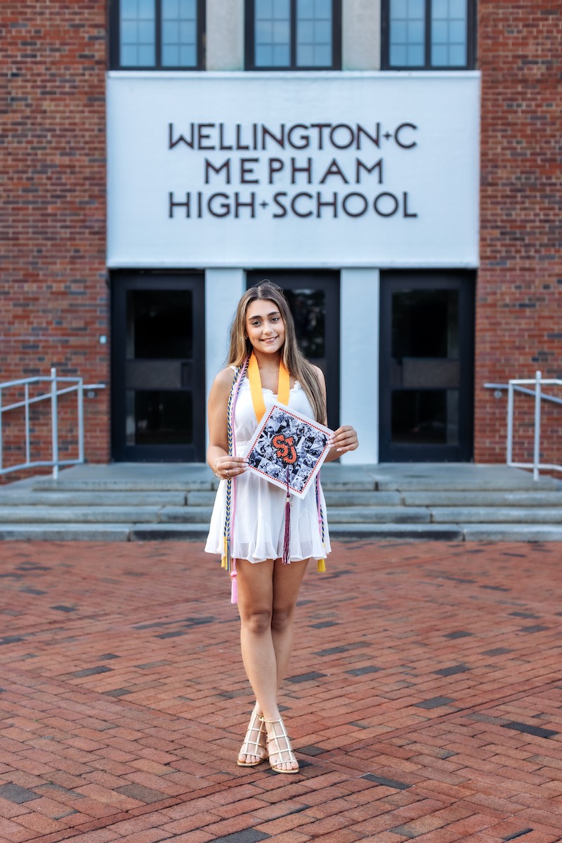 A young woman in a white dress and graduation cords stands in front of Wellington C. Mepham High School, holding a decorated graduation cap.
