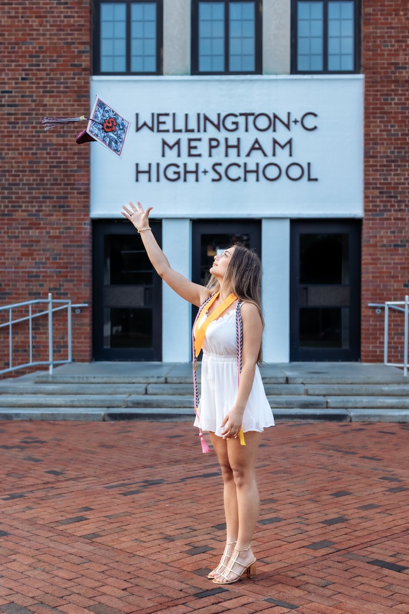 A graduate in a white dress throws her decorated cap in the air outside Wellington C. Mepham High School.