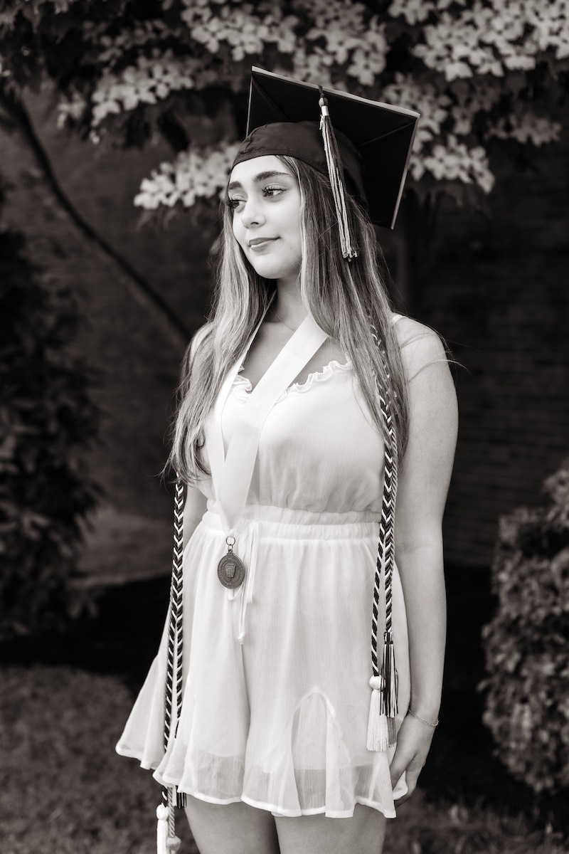 A young woman in a graduation cap and gown stands outdoors, wearing honor cords and a medal, with a neutral expression.