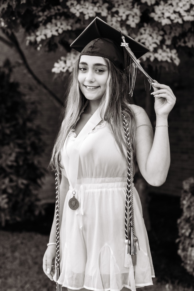 Young woman in graduation cap and gown holding tassel, wearing an honor cord and medal, standing outdoors and smiling at the camera.