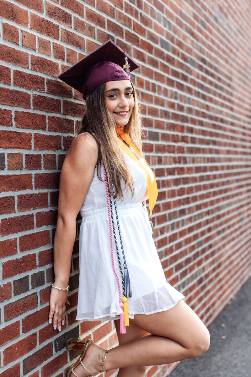 A young woman in a white dress and maroon graduation cap leans against a brick wall, wearing honor cords and a gold stole, smiling at the camera.
