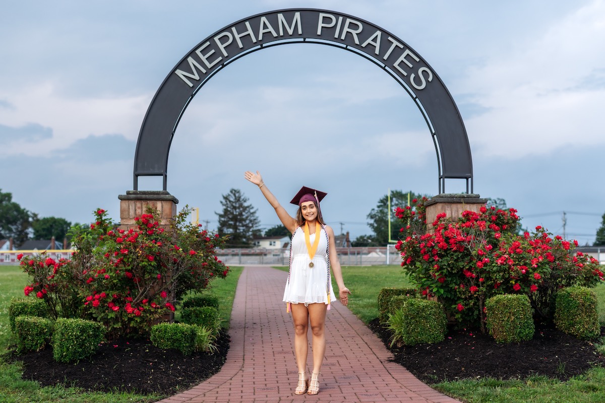 A graduate in a white dress and cap stands under a large "Mepham Pirates" arch, waving, with red flower bushes and greenery on either side.