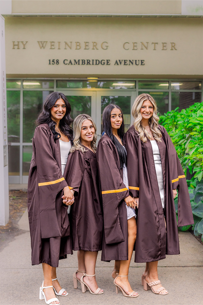 Four women in brown graduation gowns stand holding hands and smiling in front of the HY WEINBERG CENTER, 158 CAMBRIDGE AVENUE, beautifully captured by a Long Island photographer.