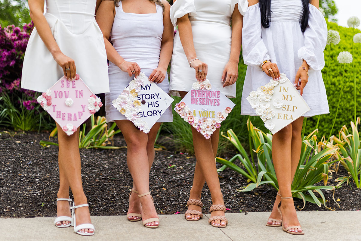 Four women in white dresses stand outdoors, holding decorated graduation caps with personalized messages; only their lower bodies are visible—a candid moment beautifully captured by a Long Island photographer.