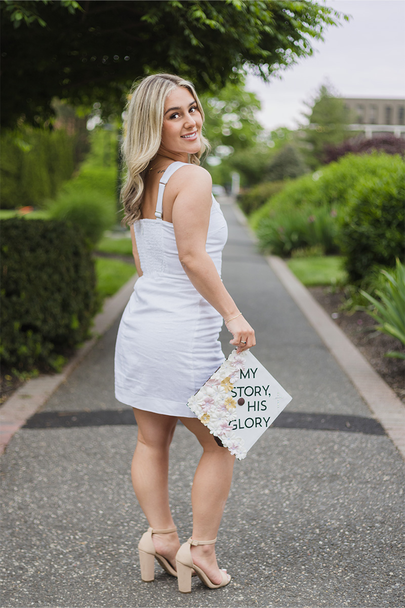 A woman in a white dress and heels stands on a path, holding a decorated graduation cap that reads "MY STORY, HIS GLORY," captured beautifully by a Long Island photographer.