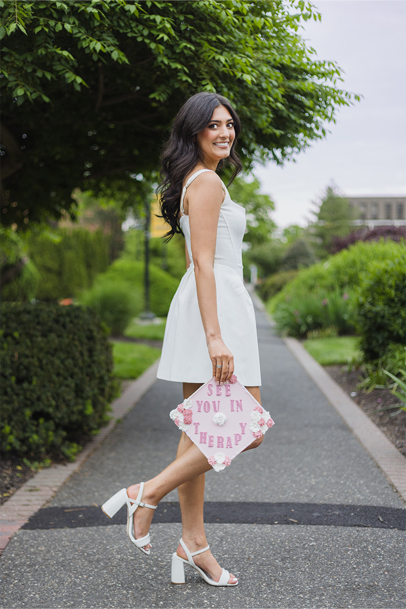 A woman in a white dress and heels stands outdoors, holding a decorated graduation cap that reads "See you in therapy," beautifully captured by a Long Island photographer.