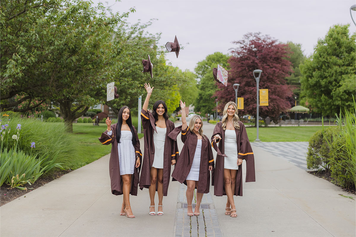 Four women in graduation gowns and white dresses walk outside, smiling and tossing their caps in the air, while a Long Island photographer captures the joyful moment among leafy trees and winding pathways.