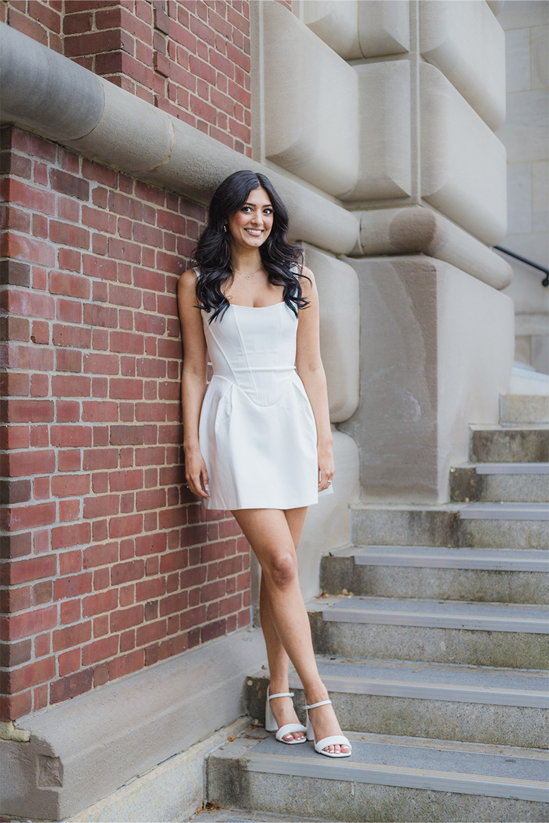 A woman in a white dress and sandals stands against a brick wall near stone steps, smiling at the camera, beautifully captured by a Long Island photographer.