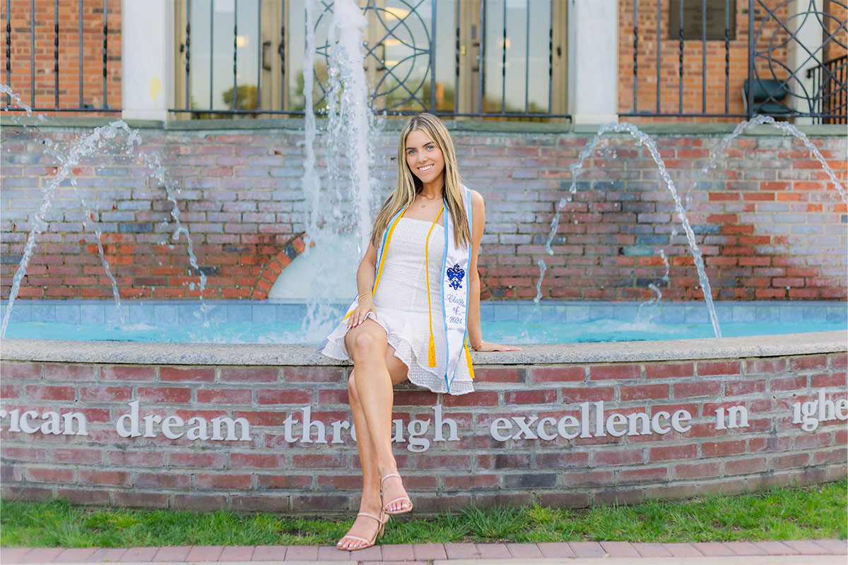 A young woman in a white dress and graduation stole sits on the edge of a fountain in front of a brick building, captured beautifully by a Long Island photographer.