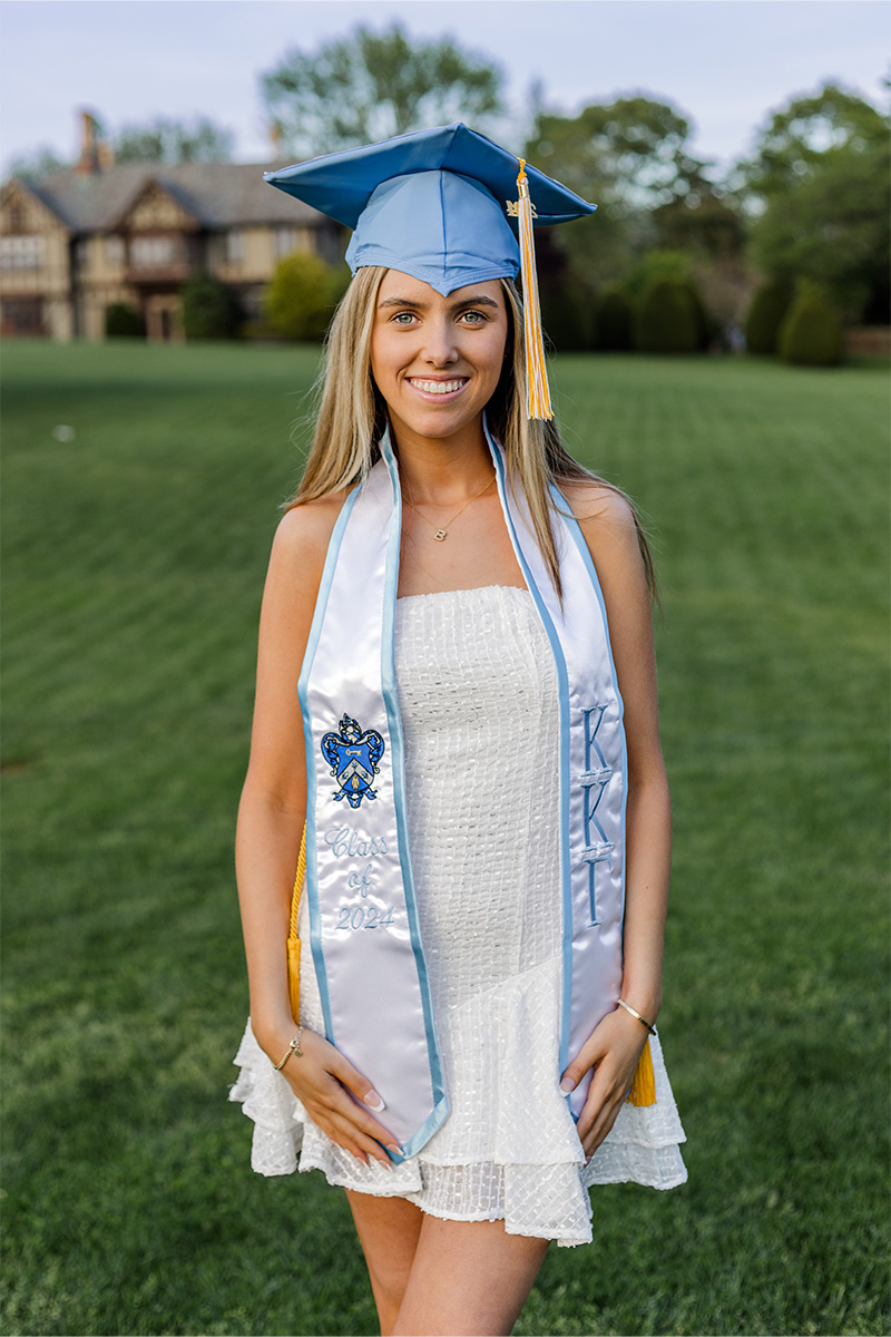 A young woman in a white dress and light blue graduation cap and stole stands on a lawn, smiling, with a building and trees in the background—captured beautifully by a Long Island photographer.