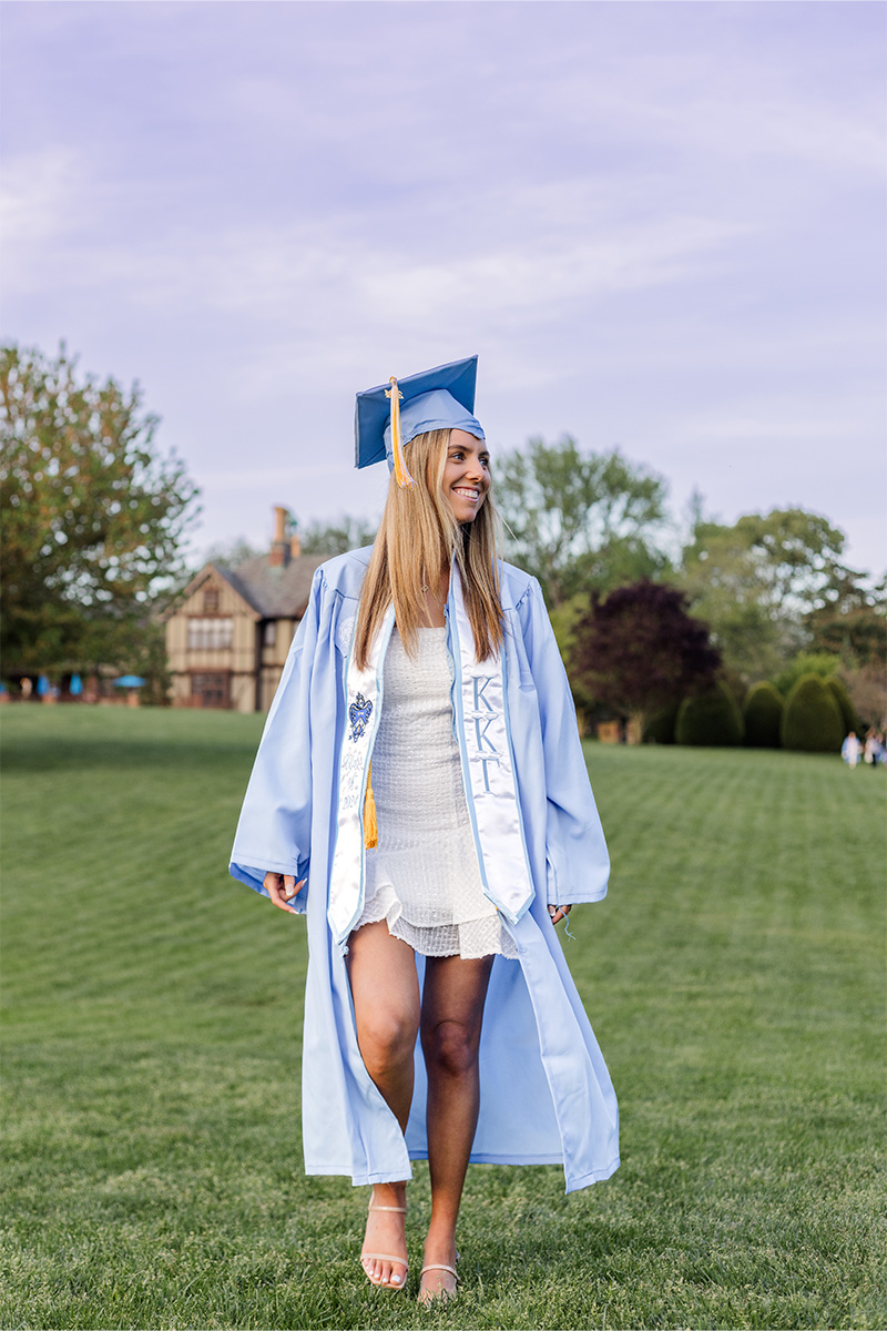 A young woman in a light blue graduation cap and gown stands on a grassy field, wearing a white dress and smiling, as captured by a Long Island photographer, with trees and a building in the background.
