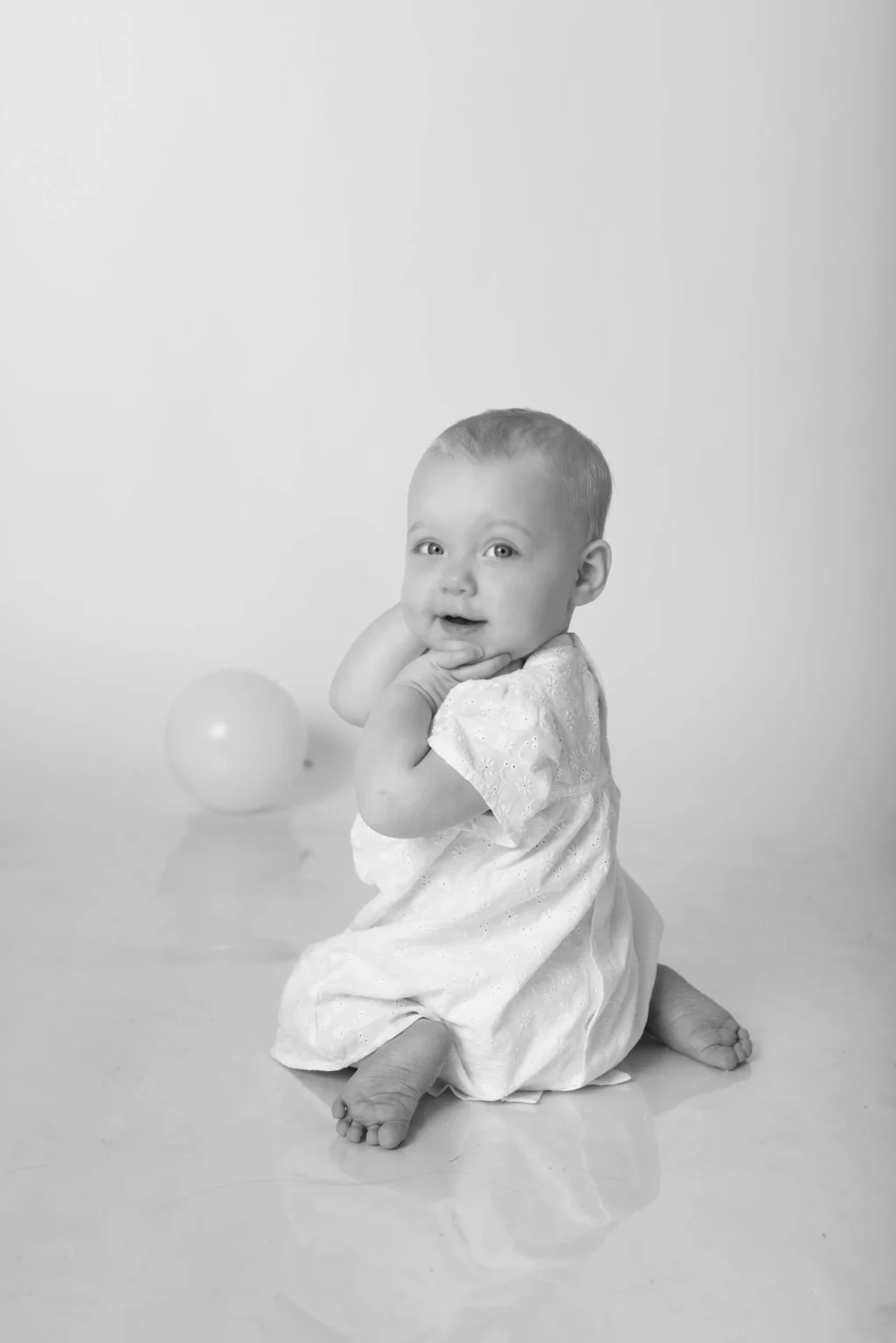 A baby in a light-colored dress sits on the floor facing backward, looking over their shoulder. A balloon is on the floor in the background. The image is in black and white.