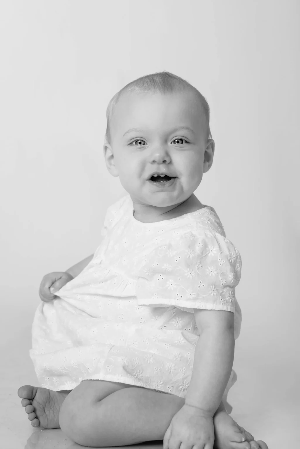 Black and white photo of a baby sitting on the floor, wearing a light-colored dress, looking at the camera and smiling with mouth slightly open.