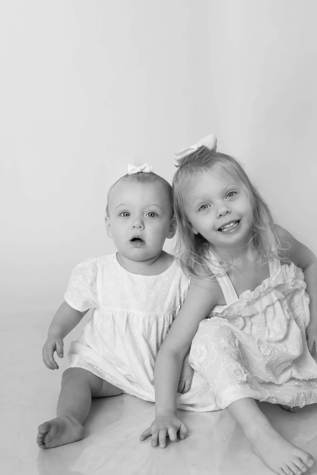 Black and white photo of two young children in light-colored dresses sitting on the floor, both with bows in their hair, facing the camera.