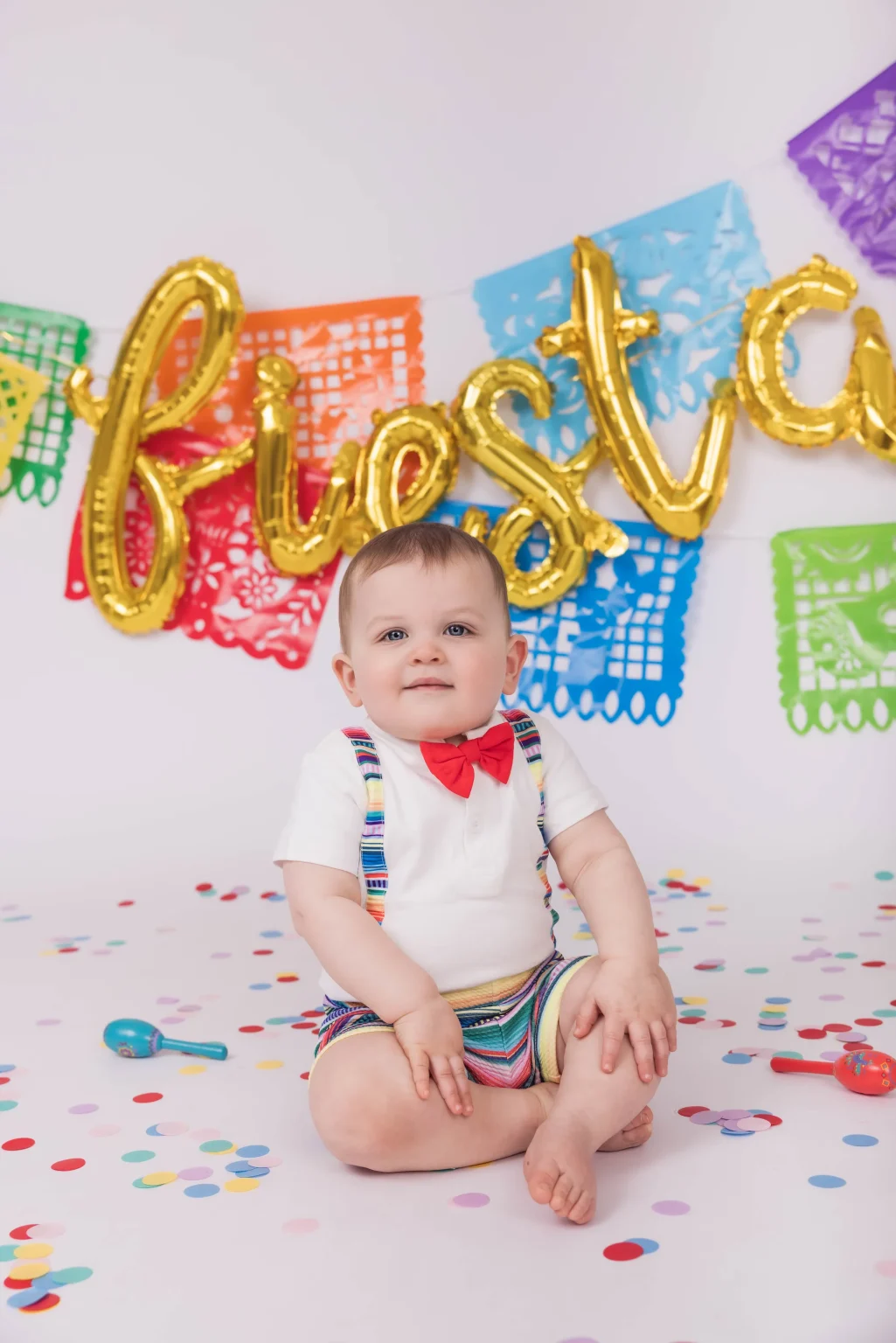 Baby sitting on floor with colorful confetti, wearing a red bow tie and rainbow suspenders, in front of a fiesta banner and papel picado decorations.