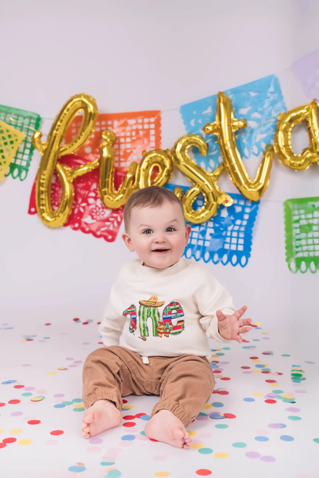Baby sitting on a confetti-covered floor, wearing a cactus-themed shirt, with colorful papel picado banners and gold "fiesta" balloon in the background.