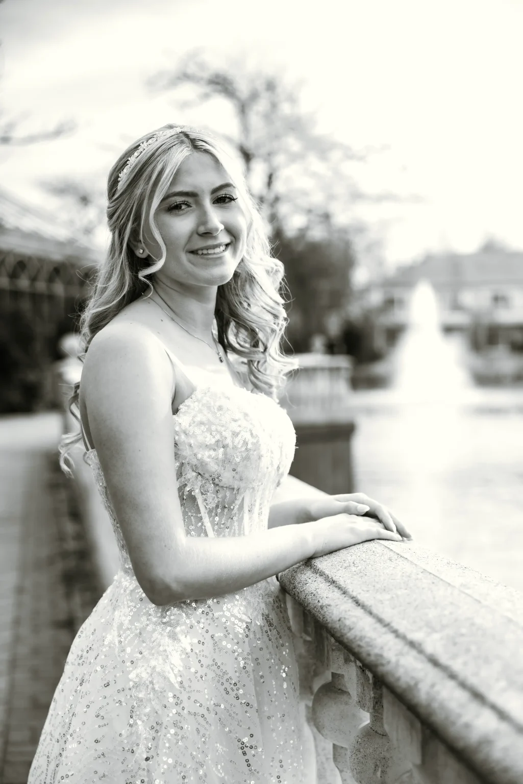 A young woman in a sequined dress stands by a stone railing, smiling at the camera in an outdoor setting with a fountain in the background.