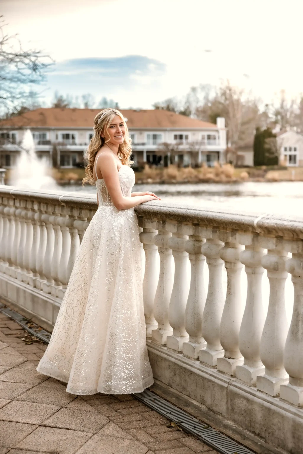 A woman in a strapless white gown stands by a white railing near a pond, with houses, trees, and a fountain in the background.