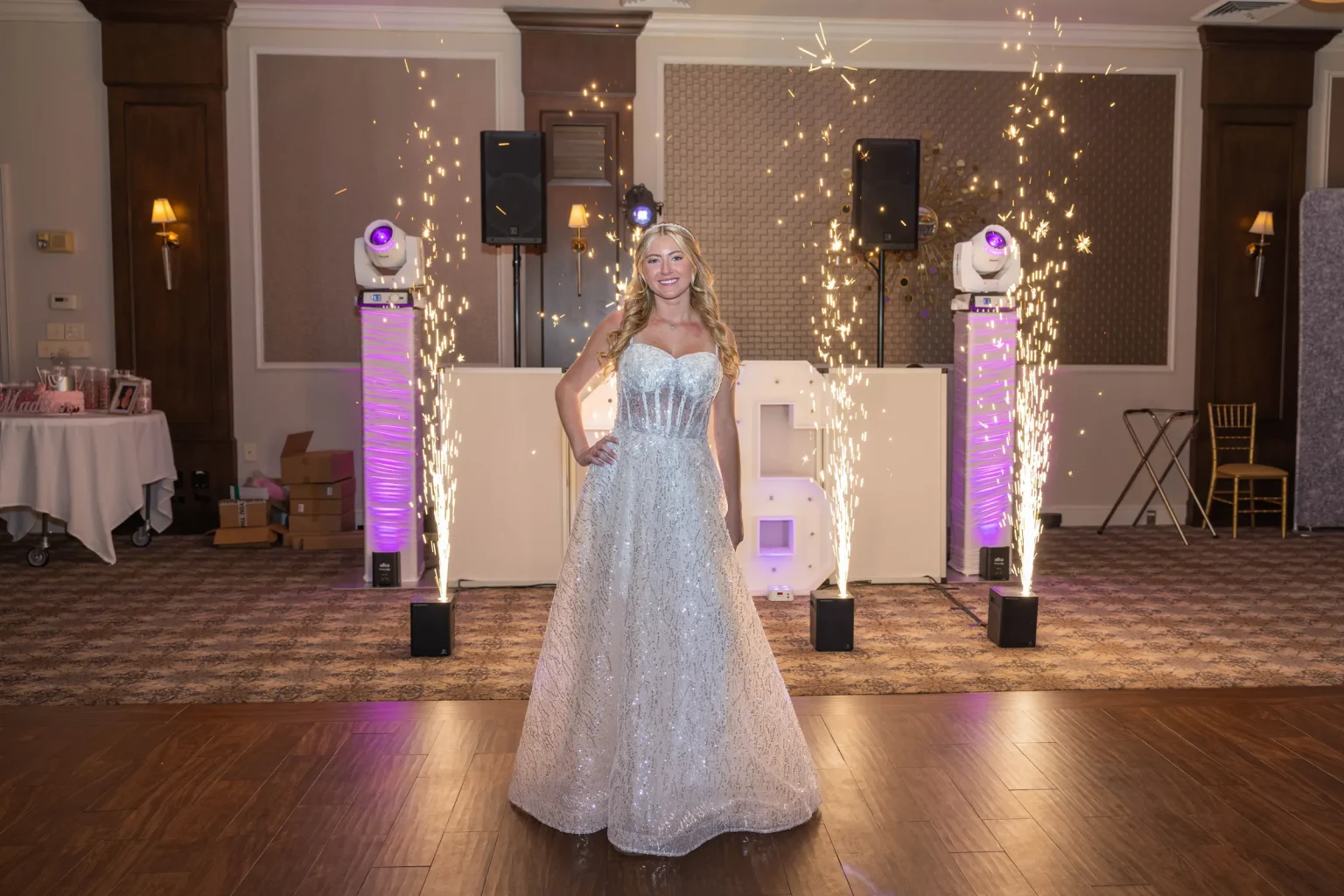A woman in a sparkling gown stands in front of illuminated decorations and sparklers at an indoor event.