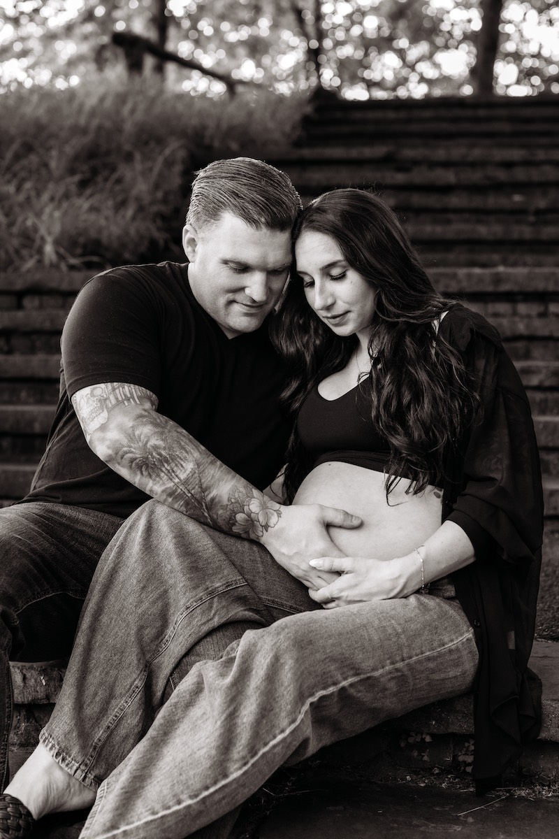 A couple sits on outdoor steps, with the man and woman both resting their hands on the woman's pregnant belly.