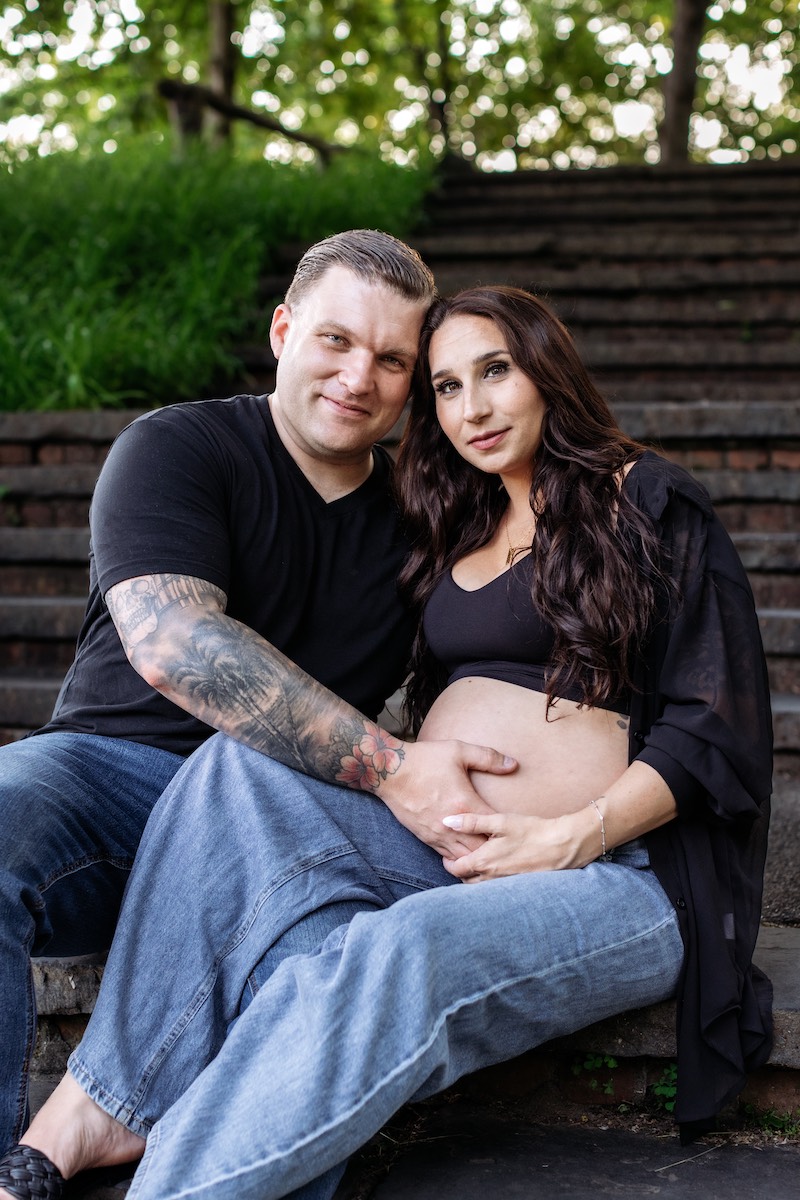 A couple sits on outdoor steps; the man has his arm around the pregnant woman and both hands rest on her exposed baby bump.