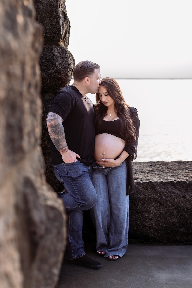 A man kisses the forehead of a pregnant woman as they stand together outside by a stone wall, with water visible in the background.