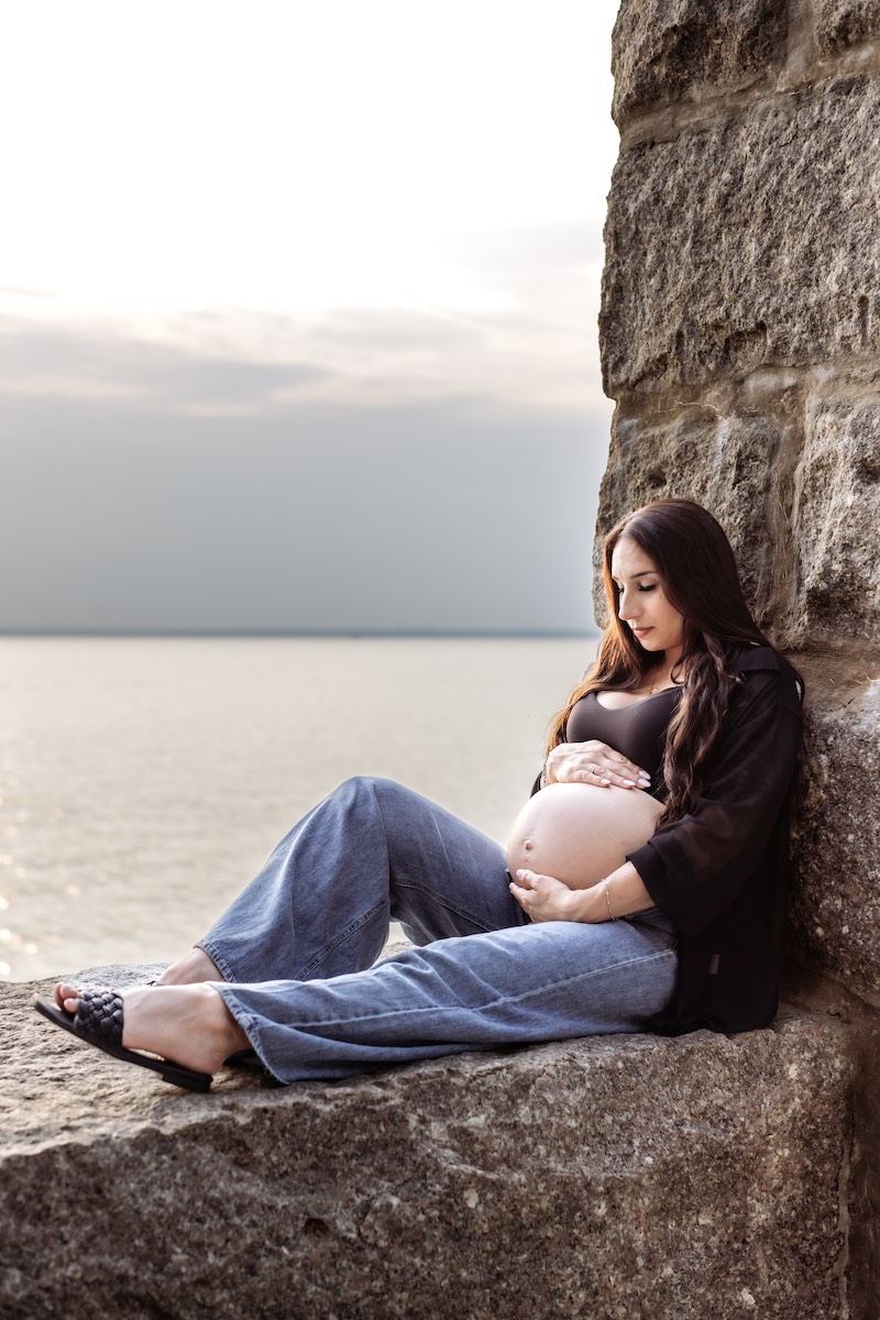 A pregnant woman sits on a stone ledge by the water, looking down and resting her hands on her belly.