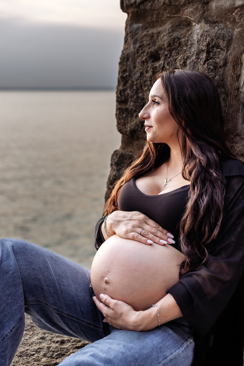 A pregnant woman with long brown hair sits by a rocky wall near water, holding her bare belly and looking into the distance.