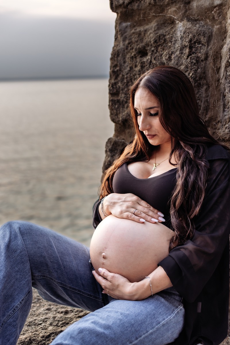 A pregnant woman sits by a stone wall near water, resting her hands on her bare belly and looking down thoughtfully.