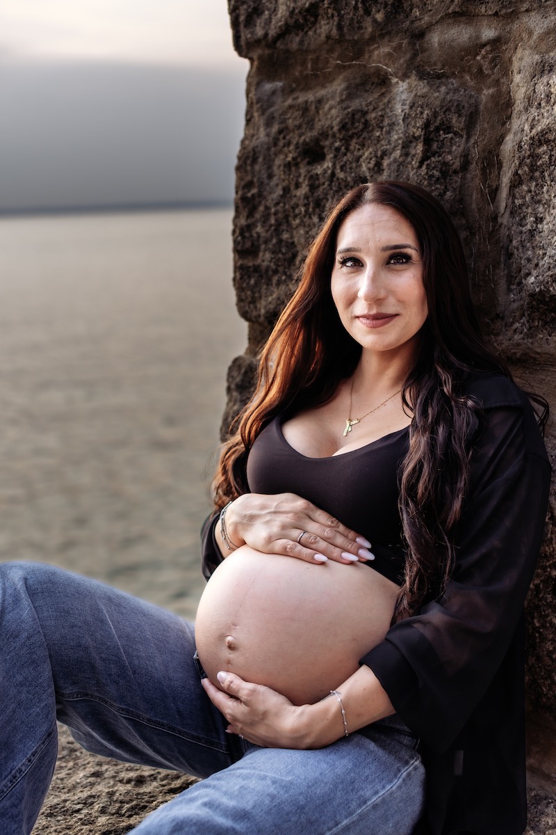 A pregnant woman with long brown hair sits outdoors by a stone wall, resting her hands on her bare belly, with water visible in the background.