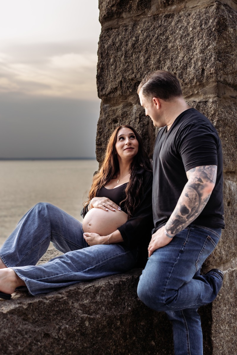 A pregnant woman sits on a stone ledge by the water, holding her belly, while a man kneels beside her, both looking at each other.
