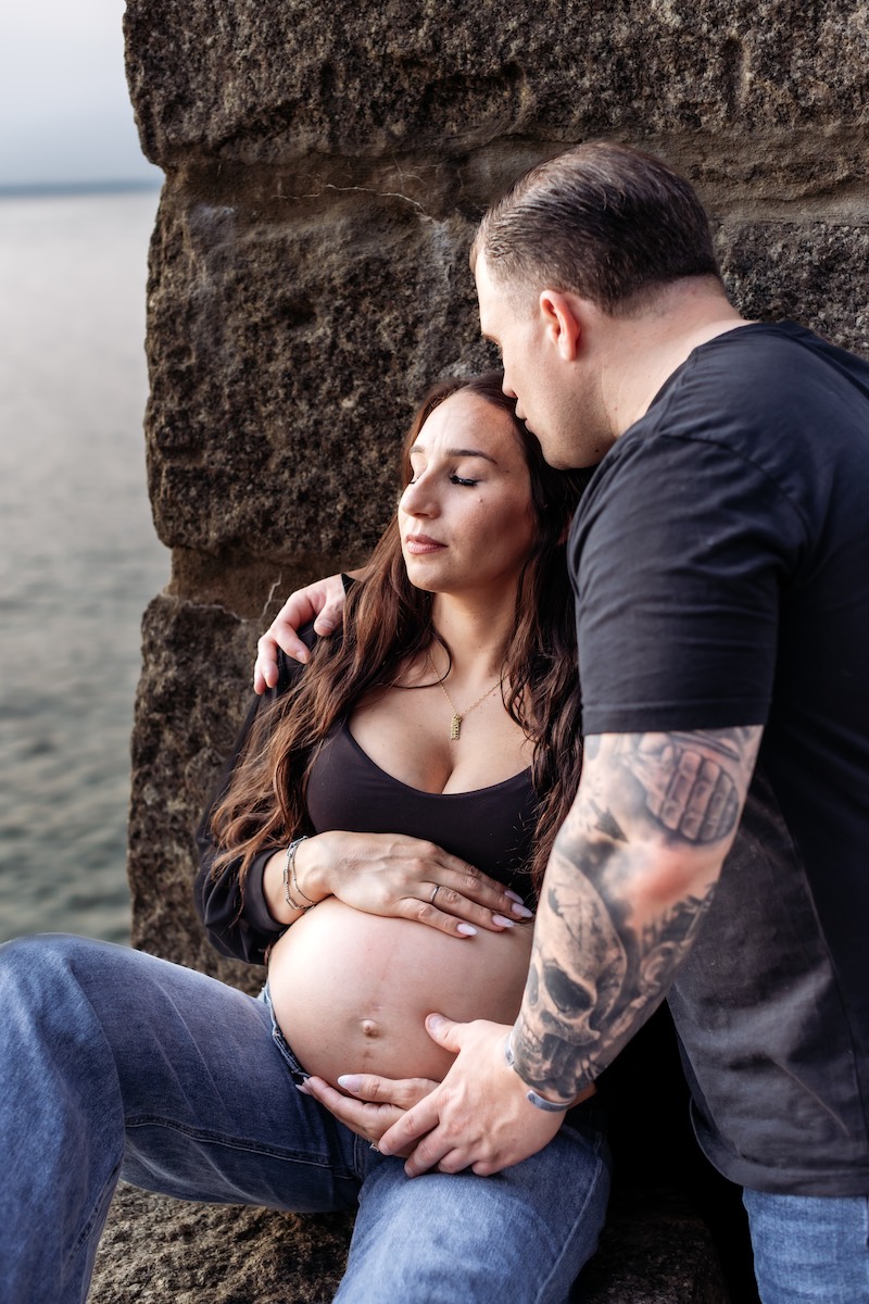 A man embraces and kisses a pregnant woman’s forehead as she rests against a stone wall by the water, with his hands on her belly.