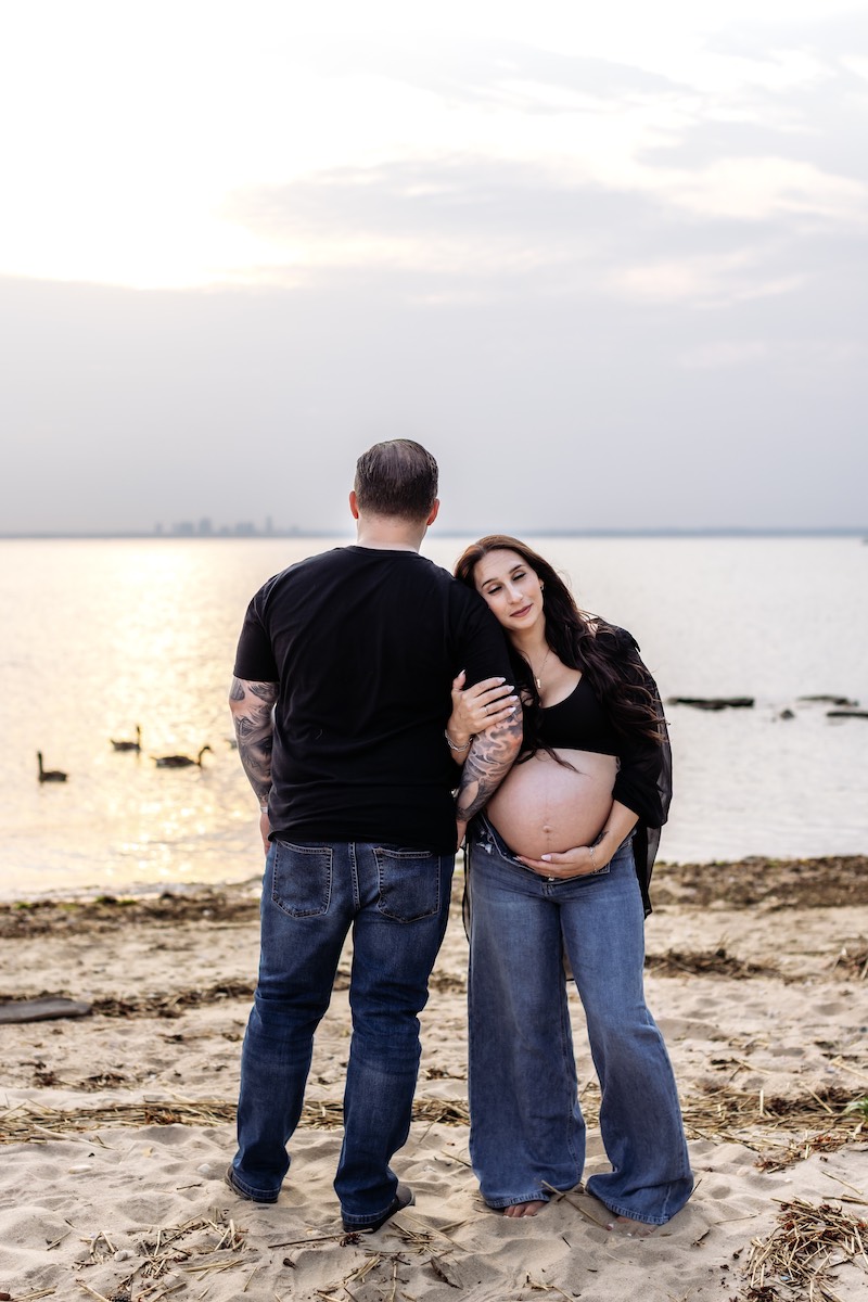 A pregnant woman stands on a beach holding her belly, leaning on a man with his back to the camera. The sun sets over the water in the background, with birds swimming nearby.