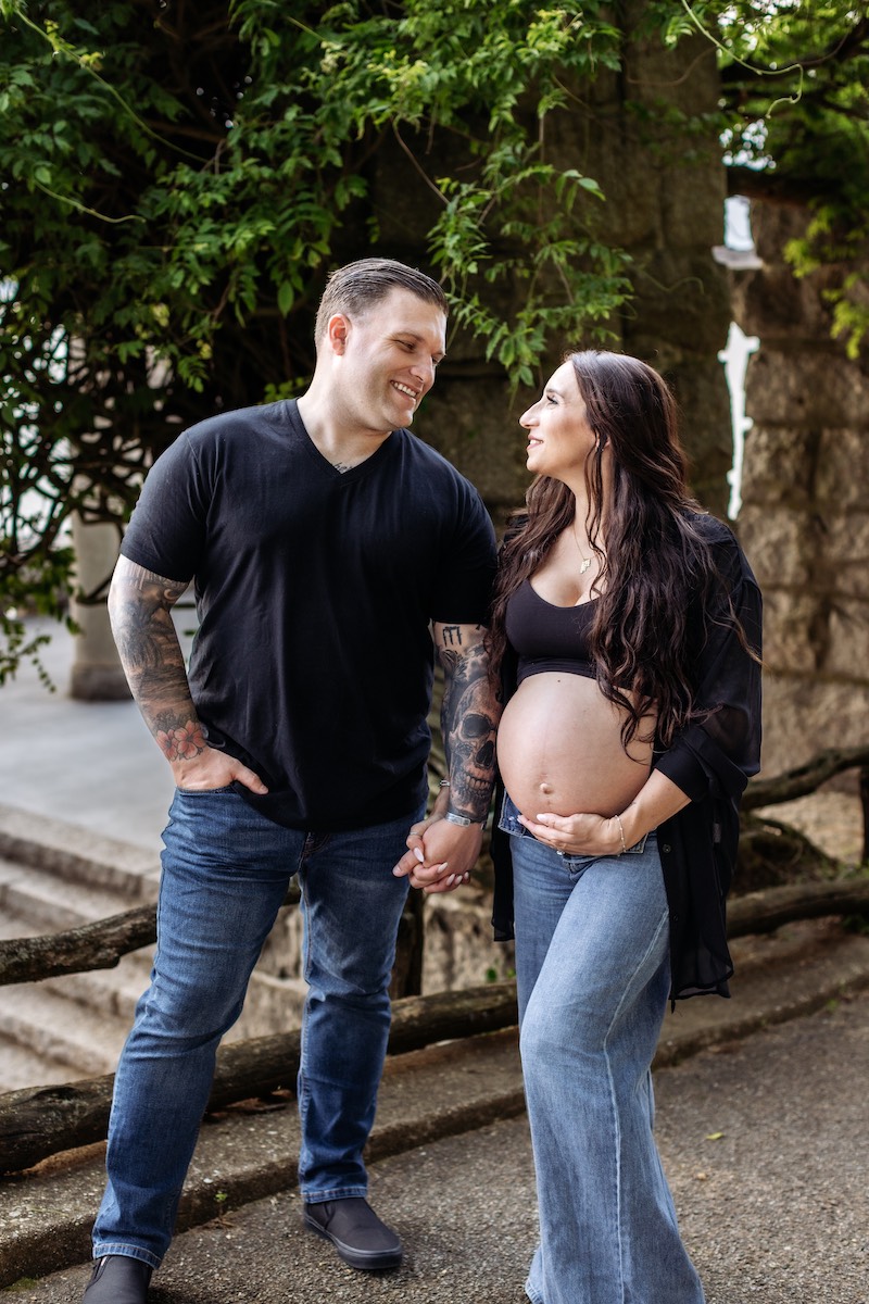 A man and a pregnant woman stand outdoors holding hands, smiling at each other. The woman has one hand on her bare belly. Both are casually dressed in jeans and black tops.