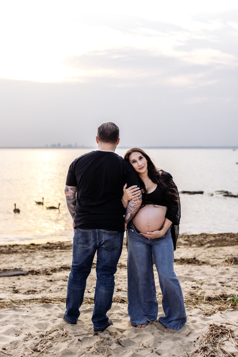 A pregnant woman stands on a sandy beach holding her belly, leaning on a man facing the water. Geese swim in the background under a cloudy sky.