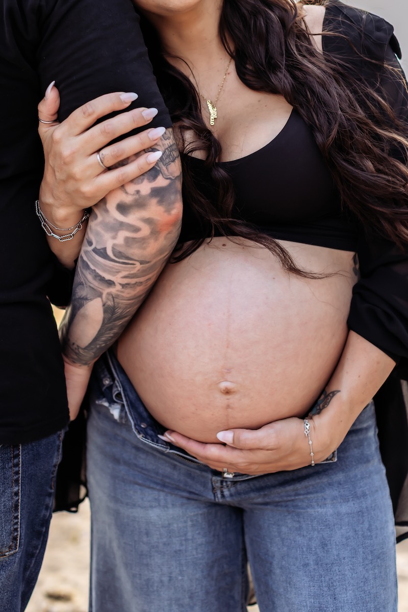 A pregnant woman in jeans and a black top holds her bare belly, while another person with tattoos stands close, their arms intertwined.
