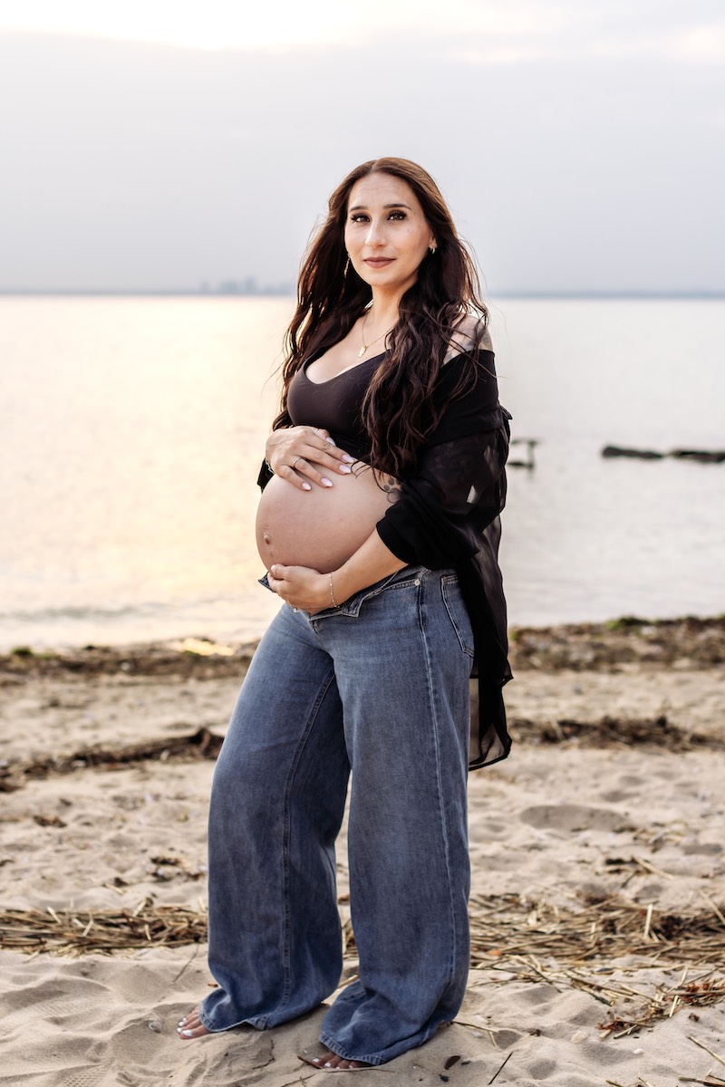 A pregnant woman stands barefoot on a sandy beach at sunset, wearing a black top, open black shawl, and loose blue jeans, with her hands resting on her belly.