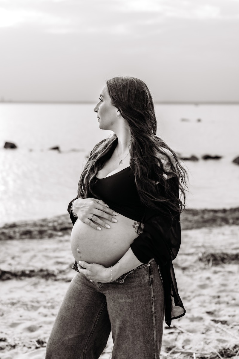 A pregnant woman stands on a beach, holding her bare belly and looking toward the water. The image is in black and white.