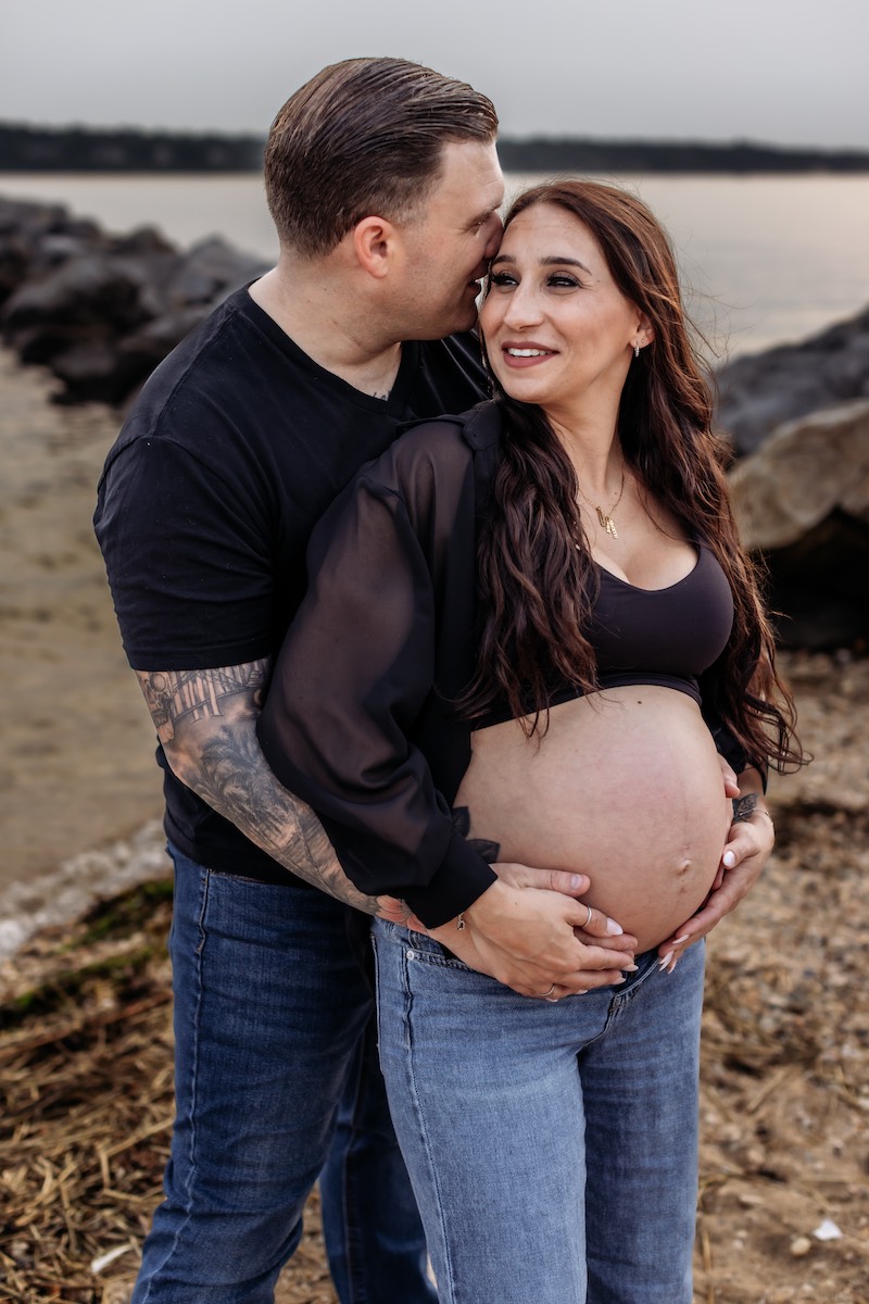 A man stands behind and kisses a pregnant woman on the forehead while holding her belly; they are outdoors near rocks and water.