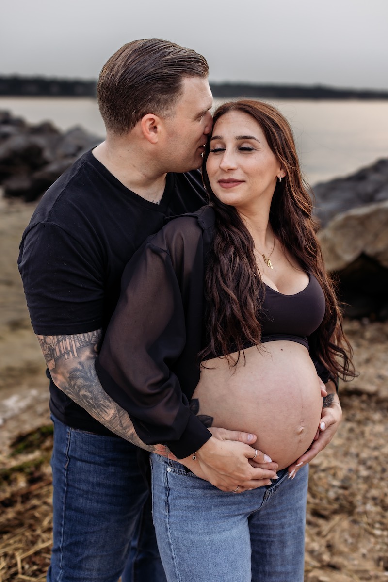 A man embraces and kisses a pregnant woman on the forehead as they stand together outdoors near water and rocks.