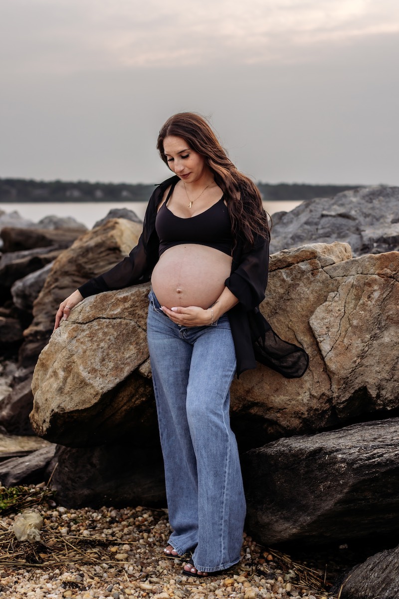 A pregnant woman in a black top and jeans stands by large rocks outdoors, gently holding her belly and looking down.