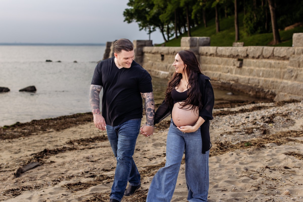 A couple walks hand-in-hand along a sandy beach; the woman is visibly pregnant and holding her belly, both are smiling at each other.