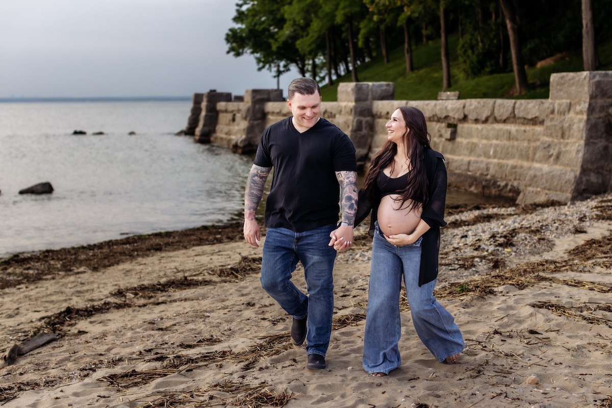 A couple walks hand in hand on a sandy beach; the woman is visibly pregnant with her shirt open, and both are smiling. Stone barriers and trees are in the background.